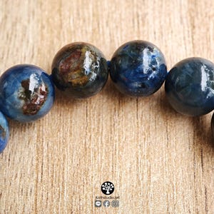 Pu&ograve; includere: Un bracciale composto da perline sferiche lucide in tonalit&agrave; blu intenso, caratterizzate da inclusioni naturali marrone scuro e nere all'interno della struttura della pietra. Le perline sono infilate su un cordino blu, con il testo Kathstudio.art visibile.