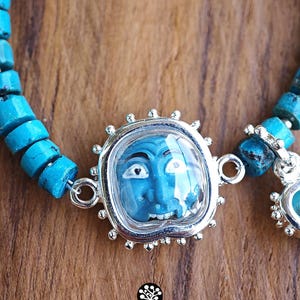 Puede incluir: Una pulsera con cuentas cil&iacute;ndricas de color turquesa y un colgante central plateado que alberga una peque&ntilde;a cara azul esculpida con detalles en blanco y negro. El colgante tiene un borde festoneado y est&aacute; unido a las cuentas por anillos.
