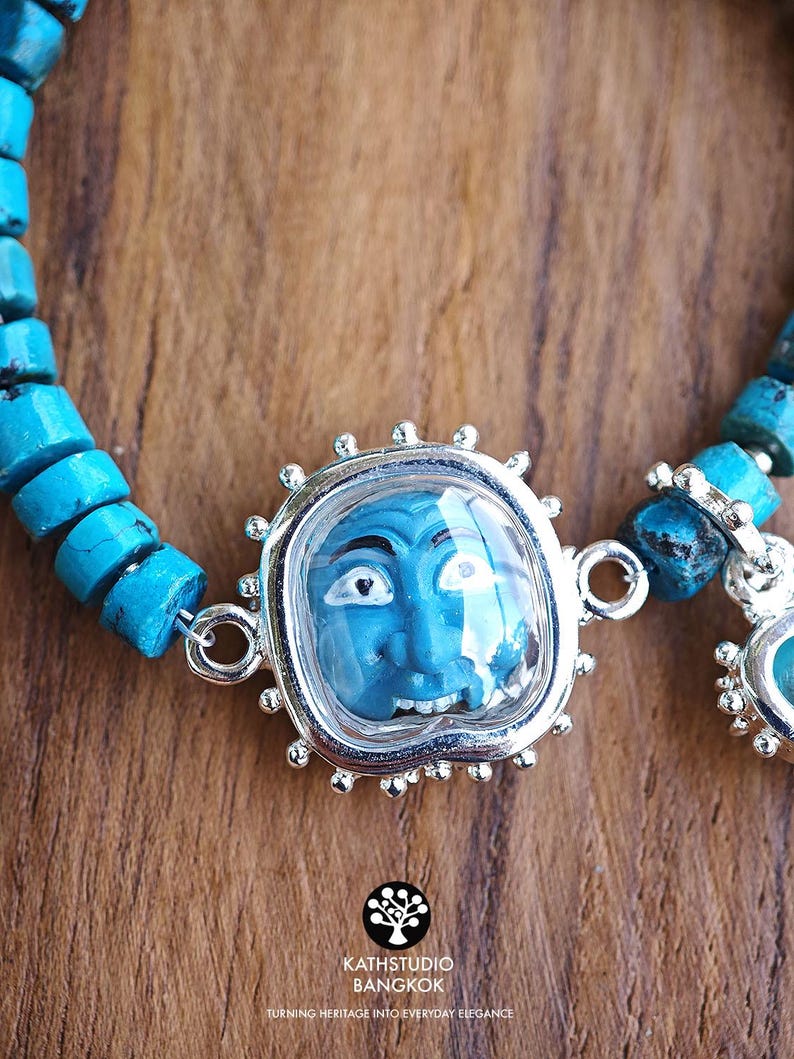 Puede incluir: Una pulsera con cuentas cil&iacute;ndricas de color turquesa y un colgante central plateado que alberga una peque&ntilde;a cara azul esculpida con detalles en blanco y negro. El colgante tiene un borde festoneado y est&aacute; unido a las cuentas por anillos.