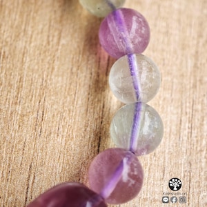 May include: A bracelet composed of spherical beads made of translucent fluorite stone, alternating between shades of pale green and deep purple. The beads are strung together, showcasing natural color variations and internal inclusions within each stone.