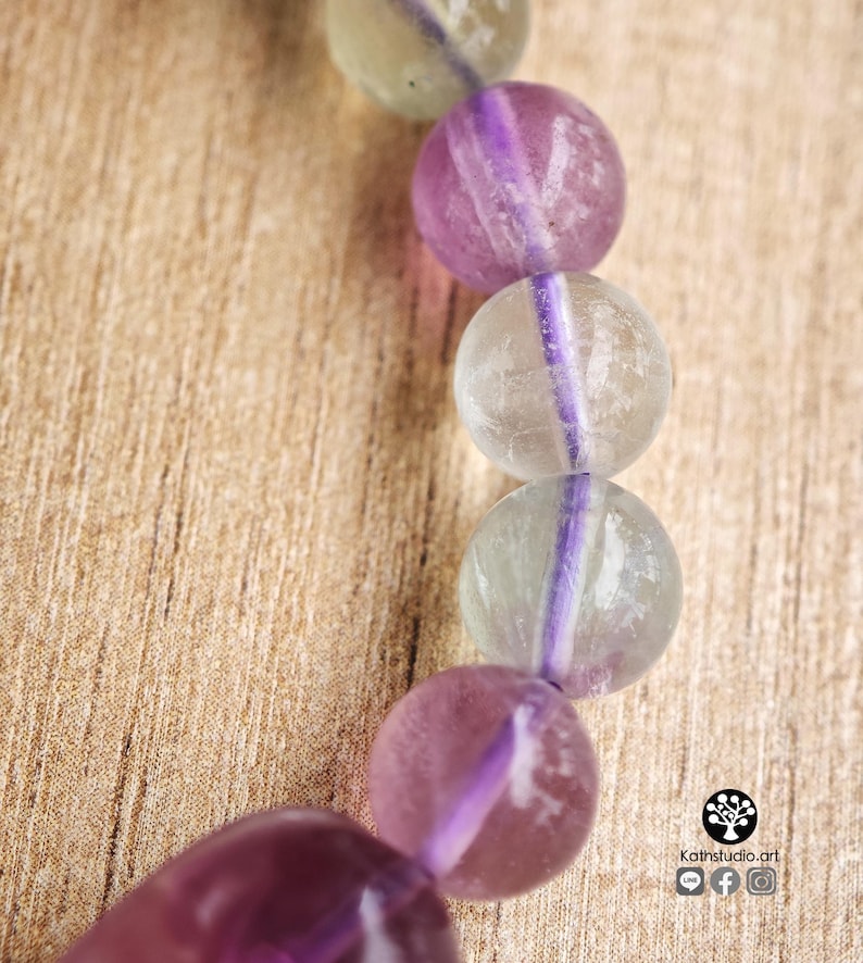 May include: A bracelet composed of spherical beads made of translucent fluorite stone, alternating between shades of pale green and deep purple. The beads are strung together, showcasing natural color variations and internal inclusions within each stone.