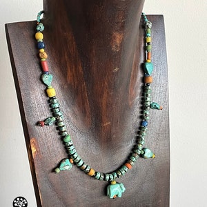 Op de afbeelding: Een turquoise kralenketting met een verscheidenheid aan kleurrijke kralen, waaronder gele, rode en blauwe. De ketting heeft een kleine turquoise olifantenhanger en wordt tentoongesteld op een houten sieradenstandaard. Een uniek sieraad.
