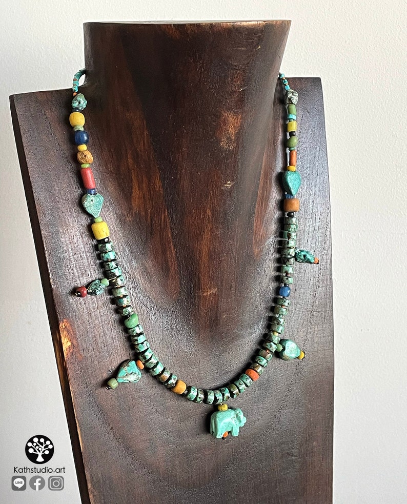 Op de afbeelding: Een turquoise kralenketting met een verscheidenheid aan kleurrijke kralen, waaronder gele, rode en blauwe. De ketting heeft een kleine turquoise olifantenhanger en wordt tentoongesteld op een houten sieradenstandaard. Een uniek sieraad.