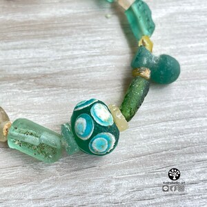 Puede incluir: Una pulsera hecha a mano con una variedad de cuentas de vidrio marino en tonos verdes, azules y crema. La cuenta focal es un &oacute;valo verde oscuro con dise&ntilde;os circulares en azul claro y blanco. La pulsera est&aacute; ensartada en un cord&oacute;n de color neutro.