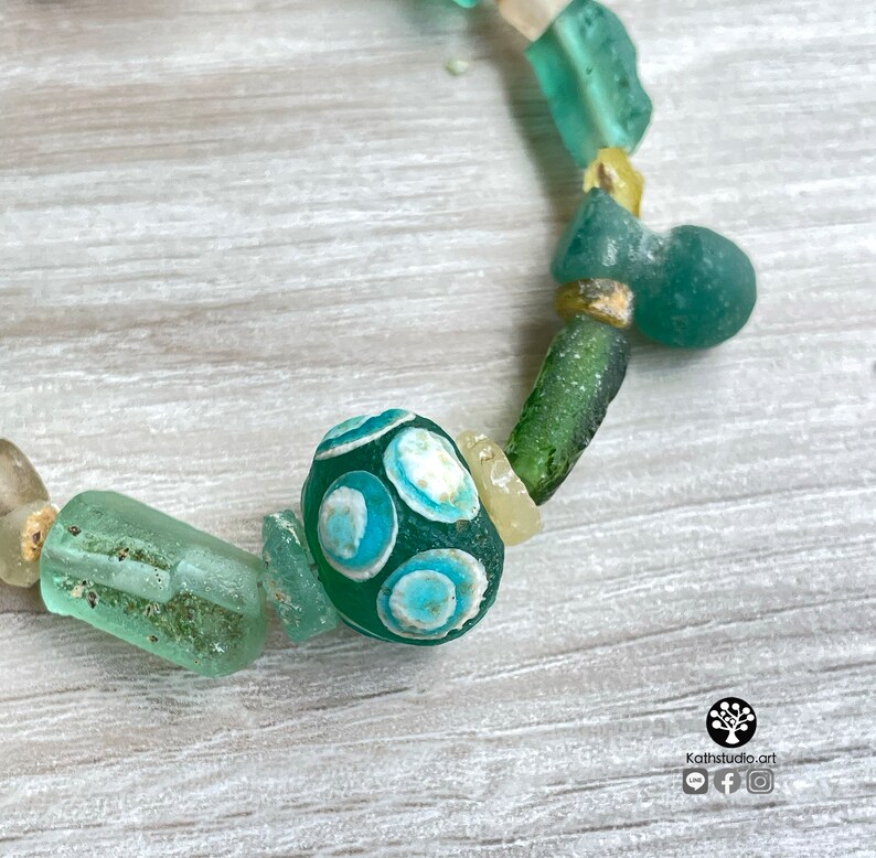 Puede incluir: Una pulsera hecha a mano con una variedad de cuentas de vidrio marino en tonos verdes, azules y crema. La cuenta focal es un &oacute;valo verde oscuro con dise&ntilde;os circulares en azul claro y blanco. La pulsera est&aacute; ensartada en un cord&oacute;n de color neutro.