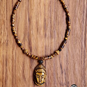 Puede incluir: Un collar que presenta una serie de cuentas cil&iacute;ndricas y esf&eacute;ricas de ojo de tigre en tonos marrones y dorados. El collar incluye un colgante central tallado con forma de cabeza de Buda, tambi&eacute;n elaborado en piedra ojo de tigre.