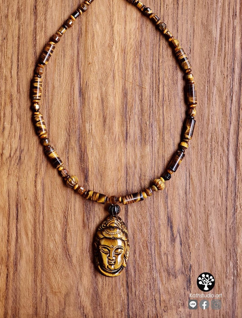 Puede incluir: Un collar que presenta una serie de cuentas cil&iacute;ndricas y esf&eacute;ricas de ojo de tigre en tonos marrones y dorados. El collar incluye un colgante central tallado con forma de cabeza de Buda, tambi&eacute;n elaborado en piedra ojo de tigre.