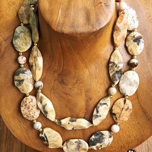 Puede incluir: Un collar de dos hilos con piedras pulidas de formas irregulares en tonos bronceado, crema, marr&oacute;n y gris. Las piedras est&aacute;n intercaladas con peque&ntilde;as cuentas redondas de color oro met&aacute;lico, creando un aspecto en capas con texturas variadas.