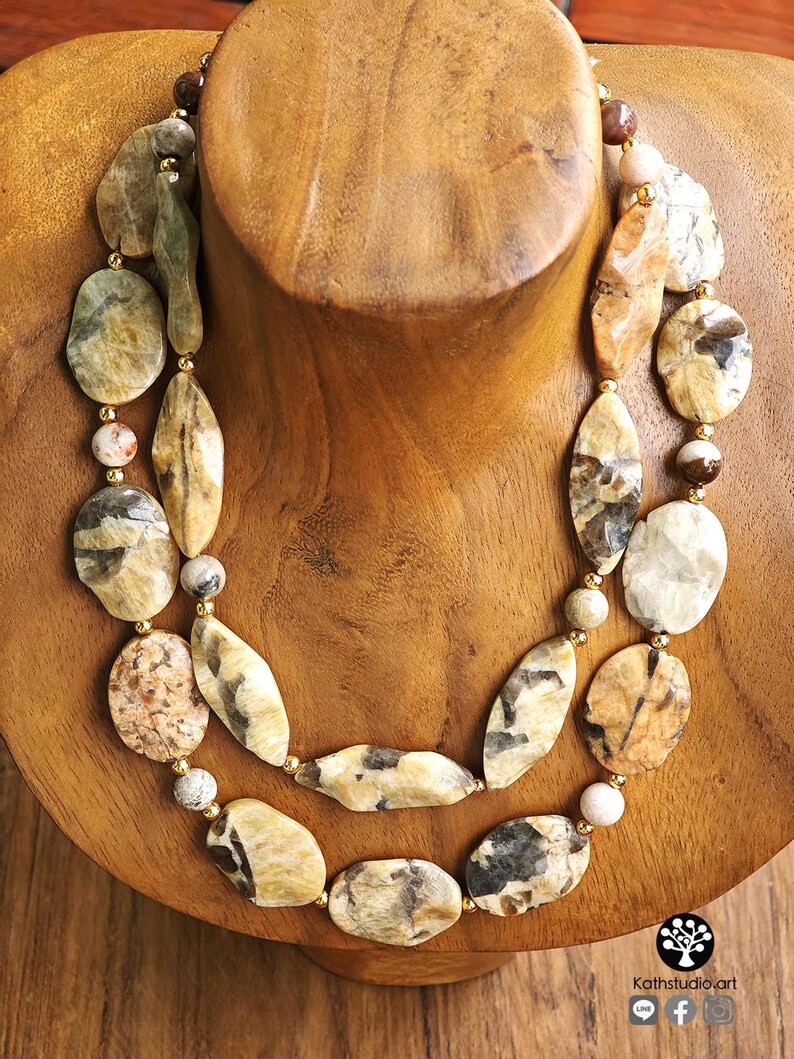 Puede incluir: Un collar de dos hilos con piedras pulidas de formas irregulares en tonos bronceado, crema, marr&oacute;n y gris. Las piedras est&aacute;n intercaladas con peque&ntilde;as cuentas redondas de color oro met&aacute;lico, creando un aspecto en capas con texturas variadas.