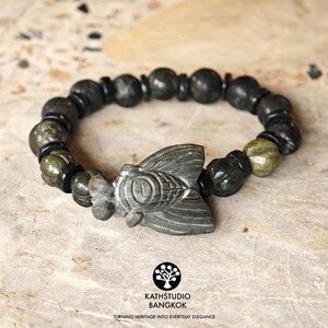 Puede incluir: Una pulsera de cuentas con un dije central de cigarra tallado en piedra gris, alternando con cuentas redondas de piedra verde oscuro y gris y peque&ntilde;os espaciadores cil&iacute;ndricos negros. La pulsera tiene una forma circular.