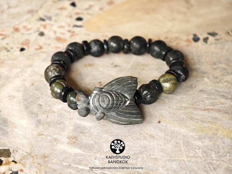 Puede incluir: Una pulsera de cuentas con un dije central de cigarra tallado en piedra gris, alternando con cuentas redondas de piedra verde oscuro y gris y peque&ntilde;os espaciadores cil&iacute;ndricos negros. La pulsera tiene una forma circular.