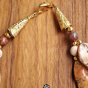 Puede incluir: Un collar con cuentas de piedra pulida en tonos marr&oacute;n y beige, intercaladas con peque&ntilde;as esferas de metal dorado. La pieza incluye terminales c&oacute;nicos ornamentados y un cierre de mosquet&oacute;n. Se observa un peque&ntilde;o logotipo con el texto Kathstudio.art.