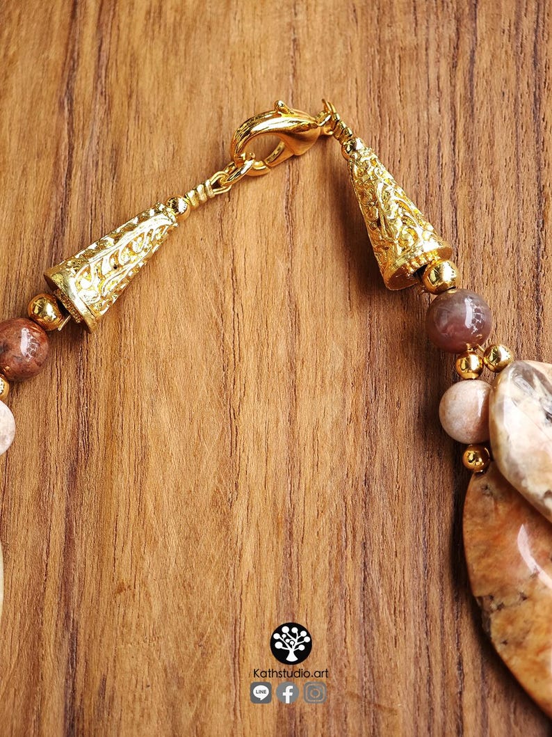 Puede incluir: Un collar con cuentas de piedra pulida en tonos marr&oacute;n y beige, intercaladas con peque&ntilde;as esferas de metal dorado. La pieza incluye terminales c&oacute;nicos ornamentados y un cierre de mosquet&oacute;n. Se observa un peque&ntilde;o logotipo con el texto Kathstudio.art.