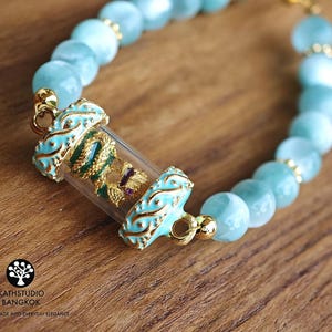 K&ouml;nnte beinhalten: Ein Armband mit hellblauen, durchscheinenden, kugelf&ouml;rmigen Perlen und einem zentralen Zylinder aus klarem Glas. Der Zylinder enth&auml;lt eine gold-gr&uuml;ne Drachenfigur mit einem kleinen violetten Akzent. Er ist mit verzierten, t&uuml;rkis emaillierten Goldmetallenden versehen.