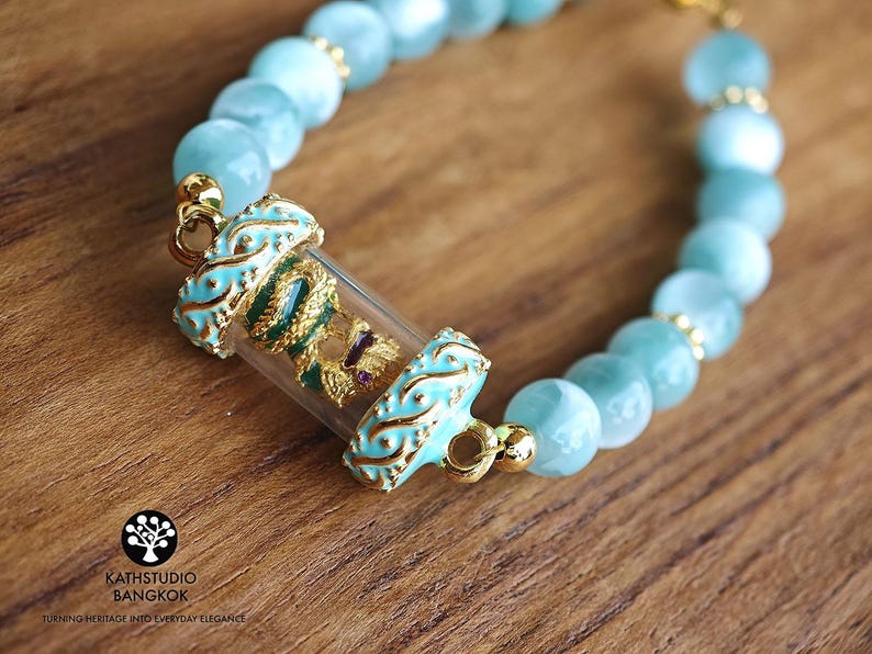 K&ouml;nnte beinhalten: Ein Armband mit hellblauen, durchscheinenden, kugelf&ouml;rmigen Perlen und einem zentralen Zylinder aus klarem Glas. Der Zylinder enth&auml;lt eine gold-gr&uuml;ne Drachenfigur mit einem kleinen violetten Akzent. Er ist mit verzierten, t&uuml;rkis emaillierten Goldmetallenden versehen.
