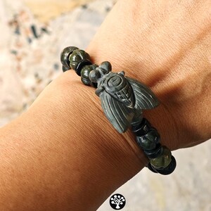 Puede incluir: Una pulsera con un dije central de cigarra tallado en piedra verde oscuro, flanqueado por cuentas redondas texturizadas de color verde oscuro y cuentas espaciadoras negras lisas. La cigarra muestra patrones de alas detallados y un cuerpo segmentado.