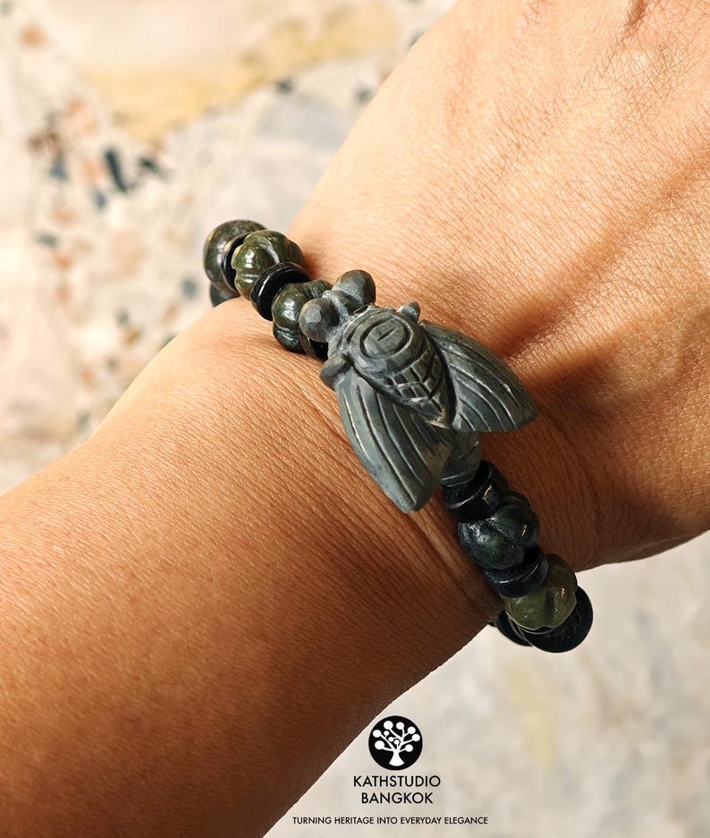 Puede incluir: Una pulsera con un dije central de cigarra tallado en piedra verde oscuro, flanqueado por cuentas redondas texturizadas de color verde oscuro y cuentas espaciadoras negras lisas. La cigarra muestra patrones de alas detallados y un cuerpo segmentado.