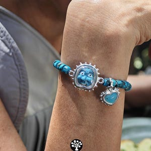 Puede incluir: Una pulsera con cuentas de turquesa azul, un marco central plateado que contiene una cara azul tallada y un dije de coraz&oacute;n plateado colgante con un centro azul. El logotipo KATHSTUDIO BANGKOK TURNING HERITAGE INTO EVERYDAY ELEGANCE es visible.