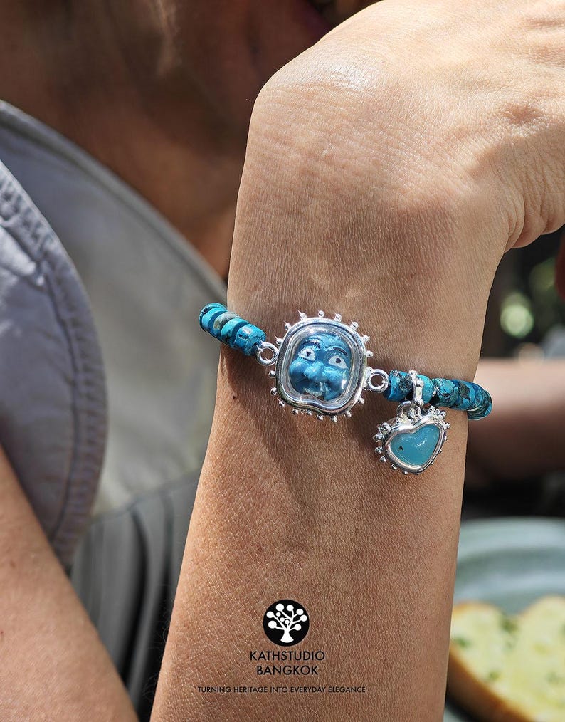 Puede incluir: Una pulsera con cuentas de turquesa azul, un marco central plateado que contiene una cara azul tallada y un dije de coraz&oacute;n plateado colgante con un centro azul. El logotipo KATHSTUDIO BANGKOK TURNING HERITAGE INTO EVERYDAY ELEGANCE es visible.