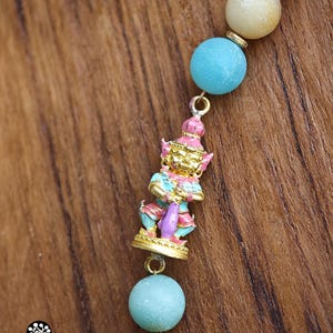 May include: A beaded necklace featuring round, matte-finish beads in shades of light blue, cream, and tan, separated by small gold-toned spacers. A central gold-toned charm depicts a standing figure with pink, blue, and gold painted details.