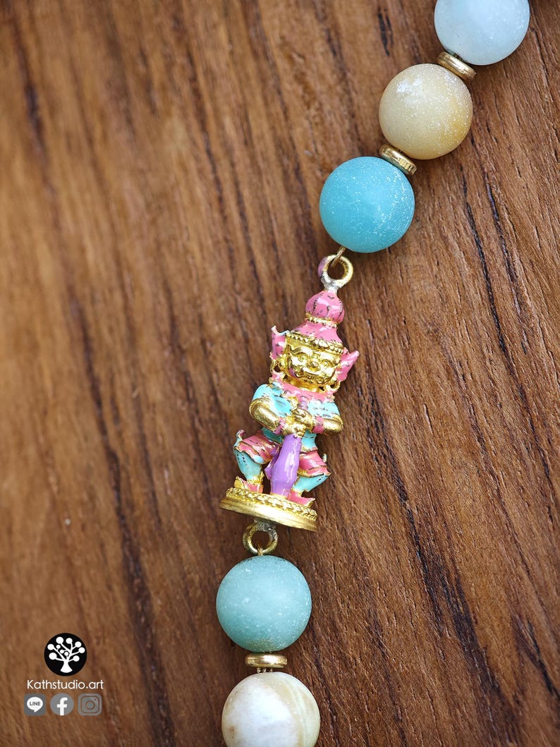 May include: A beaded necklace featuring round, matte-finish beads in shades of light blue, cream, and tan, separated by small gold-toned spacers. A central gold-toned charm depicts a standing figure with pink, blue, and gold painted details.