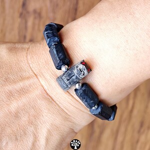 May include: A bracelet featuring black, rough-cut cylindrical stone beads interspersed with small metallic silver spheres. A central carved grey stone animal charm with red accents is positioned among the beads. Text reads Kathstudio.art.