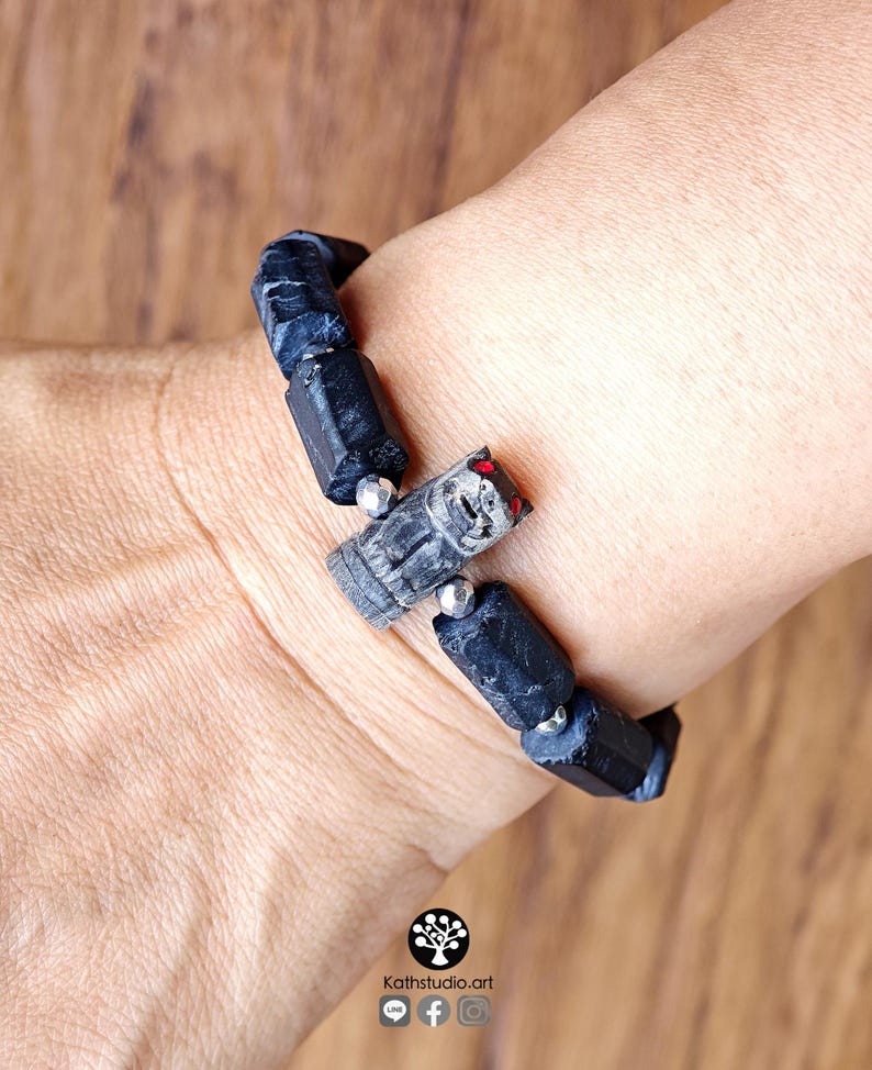 May include: A bracelet featuring black, rough-cut cylindrical stone beads interspersed with small metallic silver spheres. A central carved grey stone animal charm with red accents is positioned among the beads. Text reads Kathstudio.art.