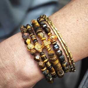 May include: A stack of bracelets featuring brown and gold tiger eye beads with faceted surfaces, interspersed with round gold-toned metal spacers and a central gold-toned cross charm, alongside two thin, textured gold-toned metal bangles.