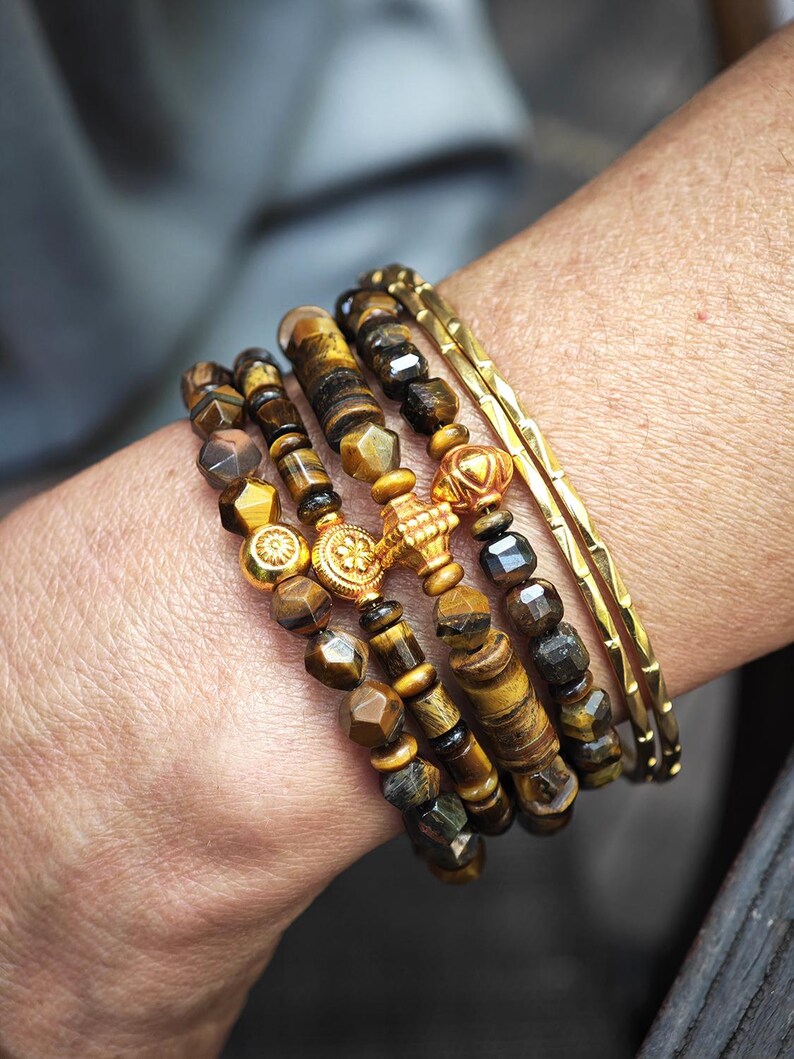 May include: A stack of bracelets featuring brown and gold tiger eye beads with faceted surfaces, interspersed with round gold-toned metal spacers and a central gold-toned cross charm, alongside two thin, textured gold-toned metal bangles.