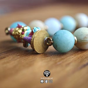 May include: A beaded bracelet featuring round, matte-finish beads in shades of light blue, cream, and tan. The bracelet includes a gold-toned metal circular clasp and a decorative charm element with small, colorful painted details in pink and blue.