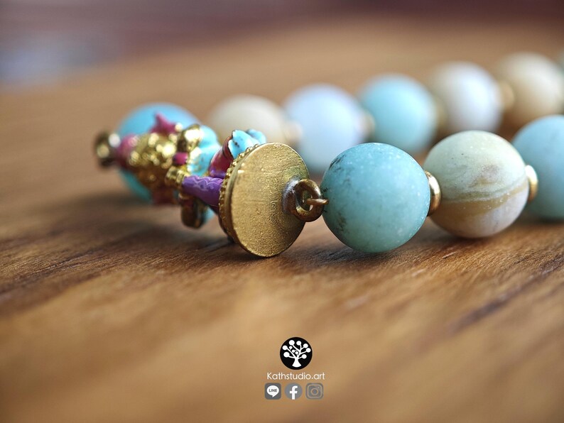 May include: A beaded bracelet featuring round, matte-finish beads in shades of light blue, cream, and tan. The bracelet includes a gold-toned metal circular clasp and a decorative charm element with small, colorful painted details in pink and blue.