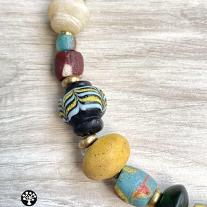 May include: A close-up of a colorful beaded necklace with a variety of shapes and sizes. The beads are made of glass and have a variety of colors, including yellow, blue, green, and brown. The necklace is strung on a cord and has a gold metal clasp.