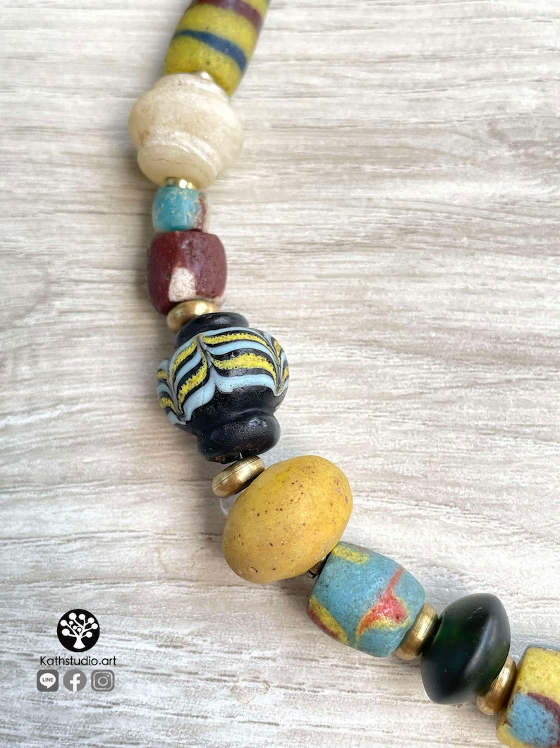 May include: A close-up of a colorful beaded necklace with a variety of shapes and sizes. The beads are made of glass and have a variety of colors, including yellow, blue, green, and brown. The necklace is strung on a cord and has a gold metal clasp.