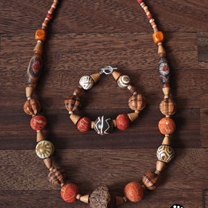 May include: A handcrafted necklace and bracelet set featuring a variety of wooden beads in shades of brown, orange, and red. The necklace and bracelet have a silver clasp. The beads have unique patterns and textures.
