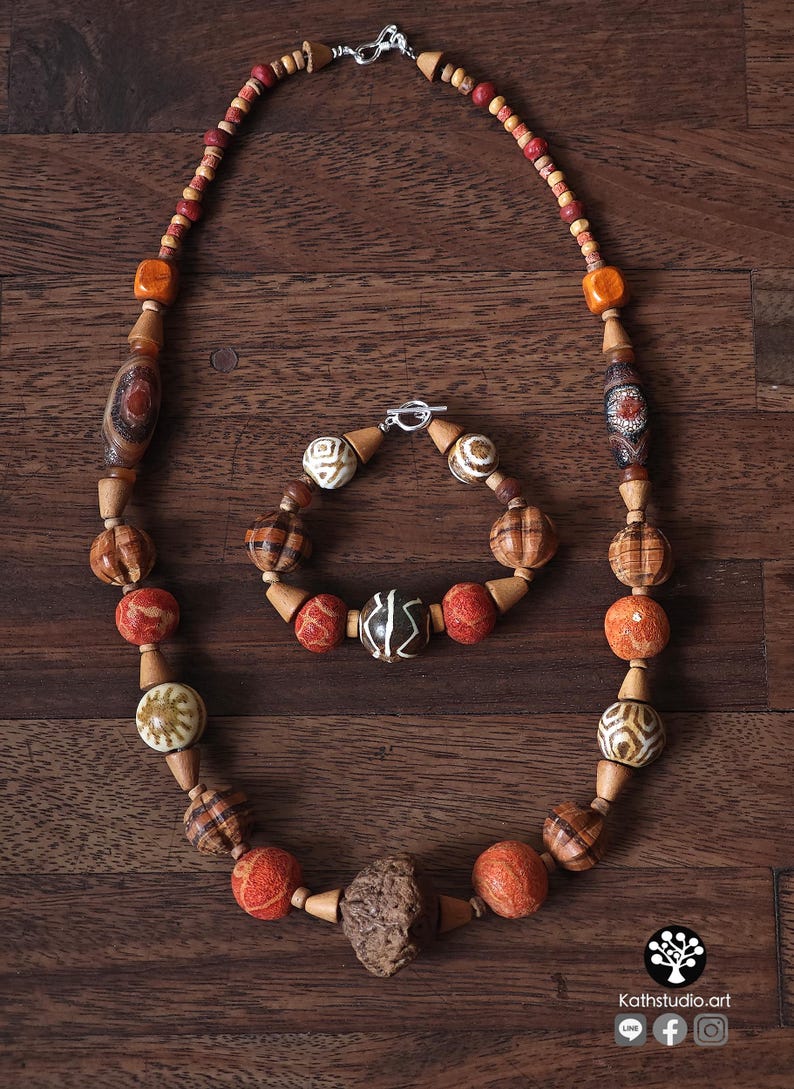 May include: A handcrafted necklace and bracelet set featuring a variety of wooden beads in shades of brown, orange, and red. The necklace and bracelet have a silver clasp. The beads have unique patterns and textures.