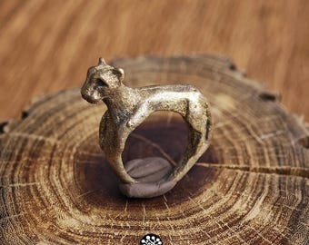 Könnte beinhalten: Ein goldfarbener Metallring in Form einer schleichenden Raubkatze. Der Ring hat eine texturierte, verwitterte Oberfläche und ein durchgehendes Banddesign. Unter dem Ring befindet sich ein kleines Logo mit dem Text KATHSTUDIO BANGKOK und einem Baum.