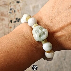 Op de afbeelding: Een elastische armband met gladde, lichtgroene en witte stenen kralen, afgewisseld met kleinere, doorschijnende gele tussenkralen. Het middelpunt is een uitgesneden, lichtgroene stenen kraal die een lachend gezicht voorstelt.