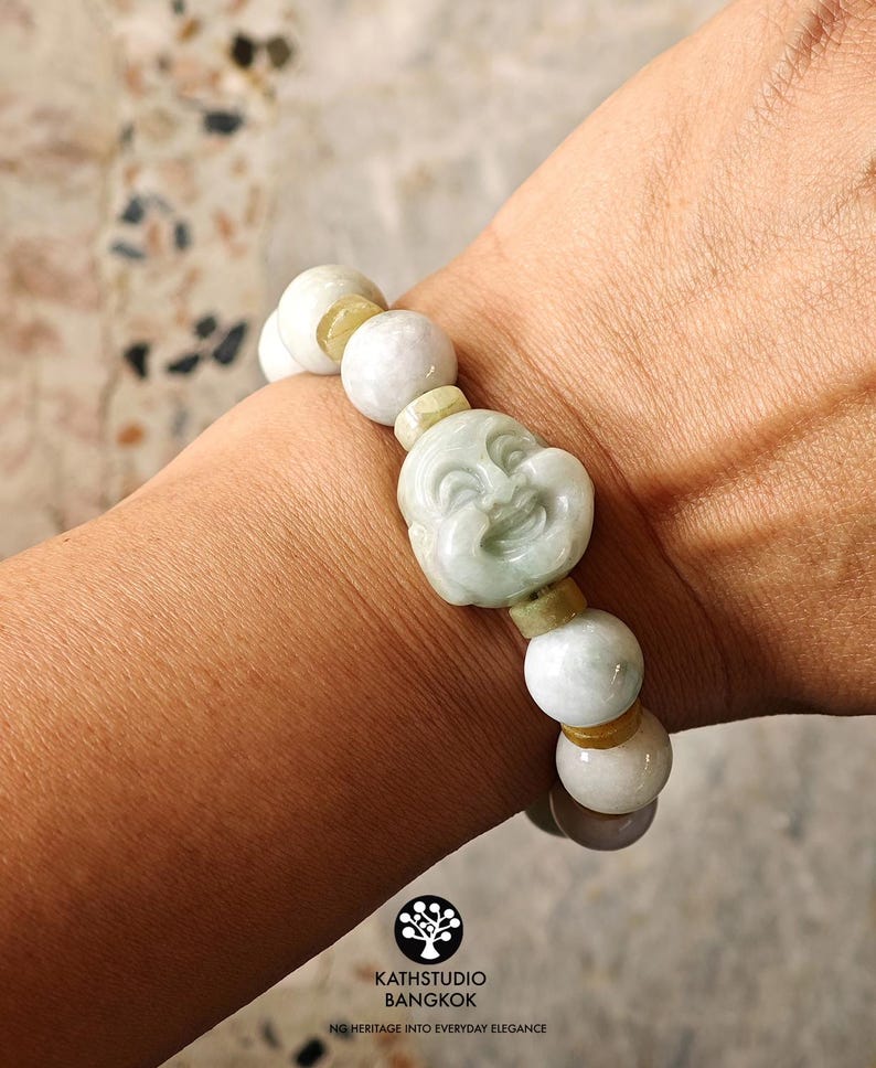 Op de afbeelding: Een elastische armband met gladde, lichtgroene en witte stenen kralen, afgewisseld met kleinere, doorschijnende gele tussenkralen. Het middelpunt is een uitgesneden, lichtgroene stenen kraal die een lachend gezicht voorstelt.