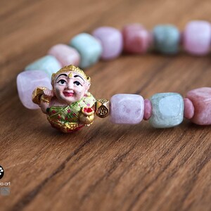 May include: A beaded bracelet featuring alternating square-cut light pink and pale green stones. A central gold-toned charm depicts a smiling figure wearing a green and gold garment, holding a small gold pot. The text Kathstudio.art is visible.