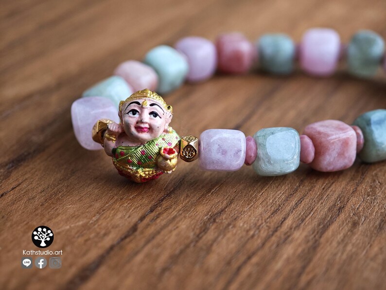May include: A beaded bracelet featuring alternating square-cut light pink and pale green stones. A central gold-toned charm depicts a smiling figure wearing a green and gold garment, holding a small gold pot. The text Kathstudio.art is visible.