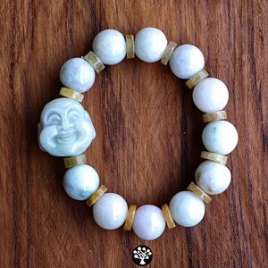 Op de afbeelding: Een kralenarmband met gladde, lichtgroene en witte stenen bollen afgewisseld met kleine, platte, amberkleurige cilindrische tussenstukjes. Een uitgesneden, lichtgroene stenen hanger die een lachend gezicht voorstelt, is in het ontwerp verwerkt.