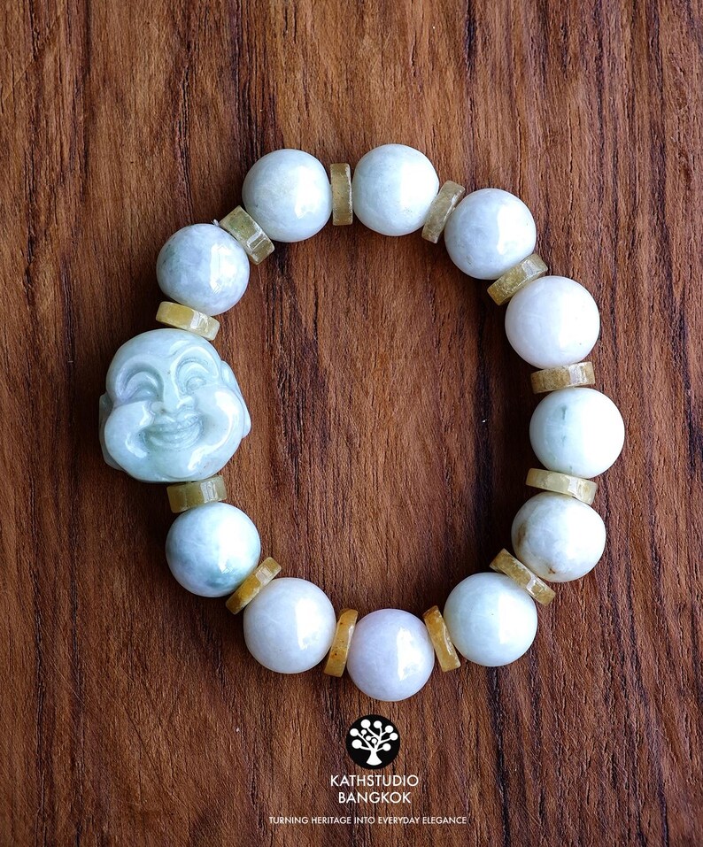 Op de afbeelding: Een kralenarmband met gladde, lichtgroene en witte stenen bollen afgewisseld met kleine, platte, amberkleurige cilindrische tussenstukjes. Een uitgesneden, lichtgroene stenen hanger die een lachend gezicht voorstelt, is in het ontwerp verwerkt.