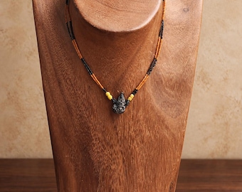 Puede incluir: Un collar con un colgante oscuro tallado suspendido de una cadena de doble hebra de pequeñas cuentas naranjas y negras, acentuado con dos cuentas amarillas en el centro. El logotipo KATHSTUDIO BANGKOK y el texto TURNING HERITAGE INTO EVERYDAY ELEGANCE aparecen debajo.