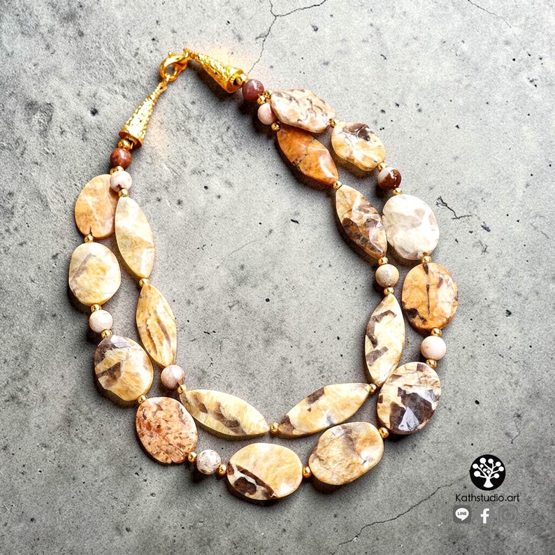 Puede incluir: Un collar de dos hilos con piedras pulidas de forma ovalada con patrones en beige y marr&oacute;n, intercaladas con peque&ntilde;as cuentas redondas de colores claros. El collar cuenta con herrajes de metal dorado y un cierre de tono dorado.