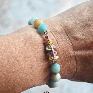 May include: A beaded bracelet featuring round, smooth, light blue and tan stones. A central gold-toned metal charm depicts a small, standing figure painted in pink, blue, and gold colors. The bracelet is strung on an elastic cord.