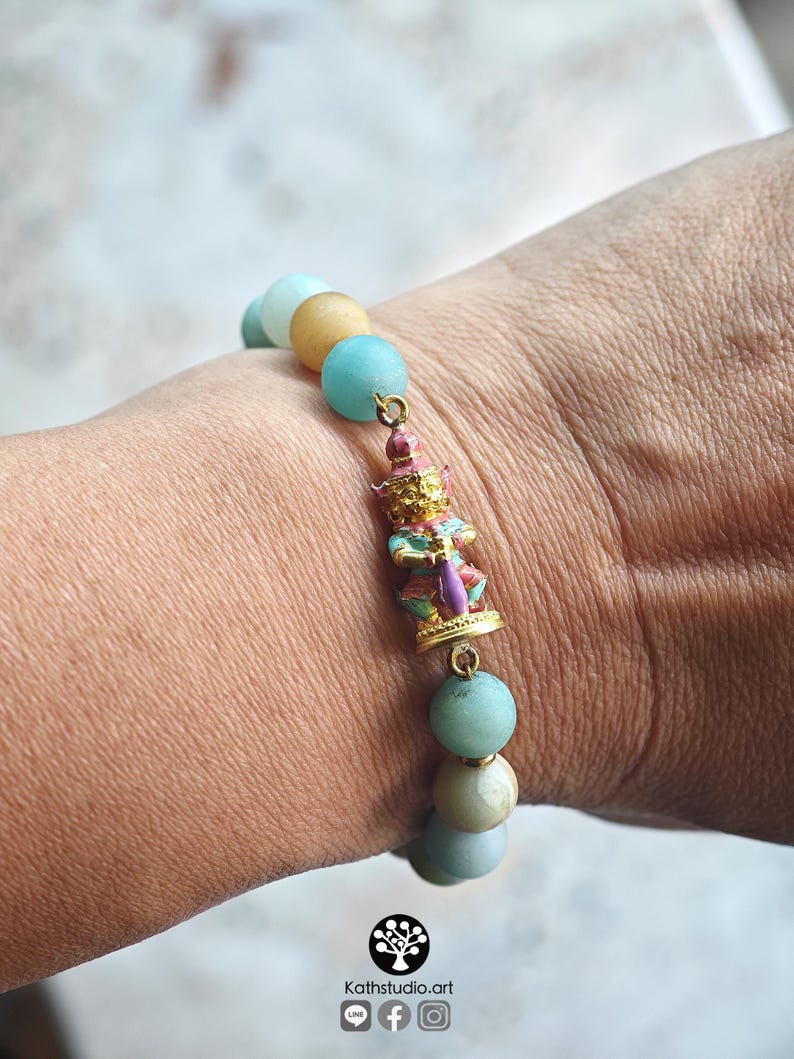 May include: A beaded bracelet featuring round, smooth, light blue and tan stones. A central gold-toned metal charm depicts a small, standing figure painted in pink, blue, and gold colors. The bracelet is strung on an elastic cord.