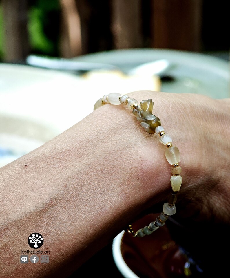 May include: A beaded bracelet featuring a mix of translucent, pale, and earthy-toned stones interspersed with small gold-colored metal spacers. The design includes a central carved stone element and a small logo with the text Kathstudio.art.