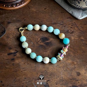 May include: A beaded bracelet featuring round, light blue and pale yellow stones interspersed with small gold-toned metal spacers. A small, detailed figurine of a deity in pink, blue, and gold tones is attached to the bracelet, which closes with a gold-toned clasp.
