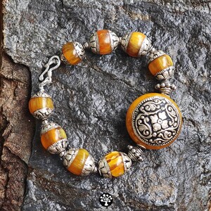 Puede incluir: Una pulsera con cuentas de color &aacute;mbar que se alternan con espaciadores de metal plateado ornamentados. Una pieza central circular grande de metal plateado con un patr&oacute;n floral en relieve est&aacute; unida a un lado, e incluye un cierre de gancho.