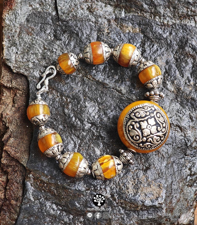 Puede incluir: Una pulsera con cuentas de color &aacute;mbar que se alternan con espaciadores de metal plateado ornamentados. Una pieza central circular grande de metal plateado con un patr&oacute;n floral en relieve est&aacute; unida a un lado, e incluye un cierre de gancho.